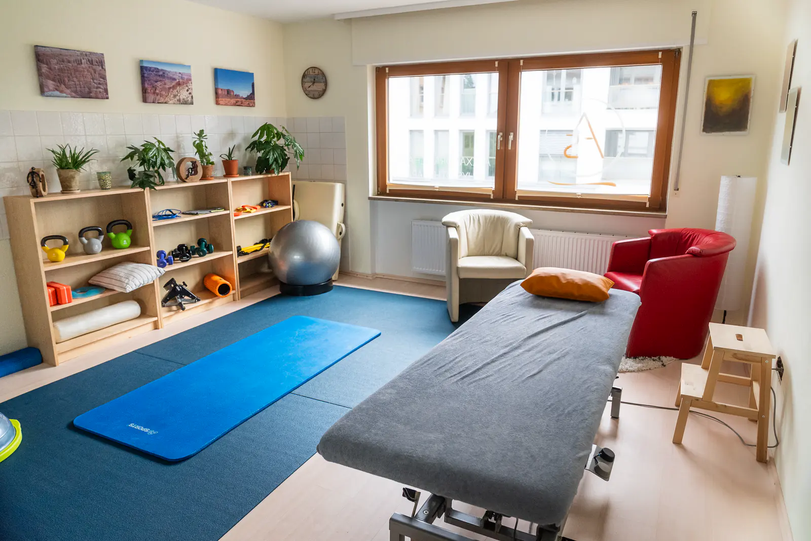 Heller Gymnastikraum mit Matte, Therapieliege, Sessel und Regal mit Kleingeräten